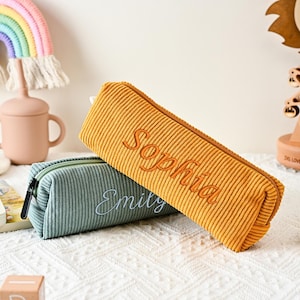 May include: Two rectangular corduroy pencil cases with zips. The mustard yellow case features the embroidered name Sophia, while the sage green case features the embroidered name Emily. Both cases have a textured ribbed fabric finish.
