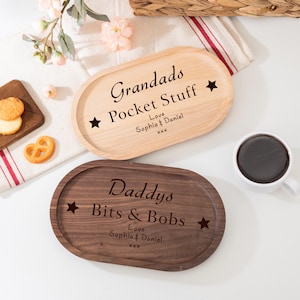 Op de afbeelding: Twee ovale houten trays, één licht en één donker, met gegraveerde tekst. De lichte tray leest "Grandads Pocket Stuff" en de donkere tray leest "Daddys Bits & Bobs". Beide bevatten namen en sterren.