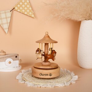 May include: A wooden carousel music box featuring four carved horses in shades of brown and white. The carousel has a light brown roof and is mounted on a round base. The name "Owen" is engraved on the base, along with a bee symbol.
