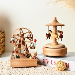 May include: A wooden ferris wheel music box featuring small animal figures in labeled gondolas A through F, alongside a wooden carousel music box with three horse figures. Both items are crafted from light-toned wood with natural grain details.