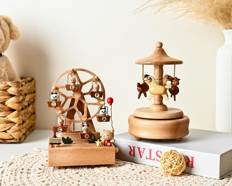 May include: A wooden ferris wheel music box featuring small animal figures in labeled gondolas A through F, alongside a wooden carousel music box with three horse figures. Both items are crafted from light-toned wood with natural grain details.