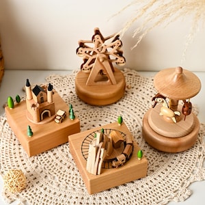 May include: Four wooden music boxes featuring miniature scenes: a castle with small trees, a ferris wheel, a train on a track with a house, and a carousel with a horse. Each item is crafted from light-toned wood with small green accents.