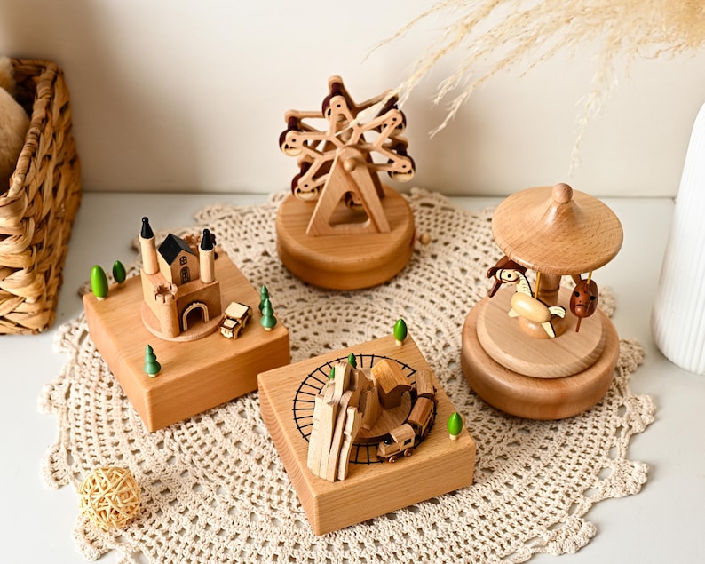May include: Four wooden music boxes featuring miniature scenes: a castle with small trees, a ferris wheel, a train on a track with a house, and a carousel with a horse. Each item is crafted from light-toned wood with small green accents.