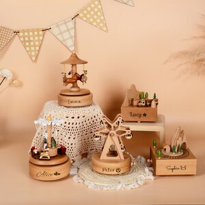 May include: A collection of wooden music boxes with various designs, including a carousel, Ferris wheel, and mountain scene. Each box is engraved with a name. The boxes are made of light-colored wood and sit on a neutral background.