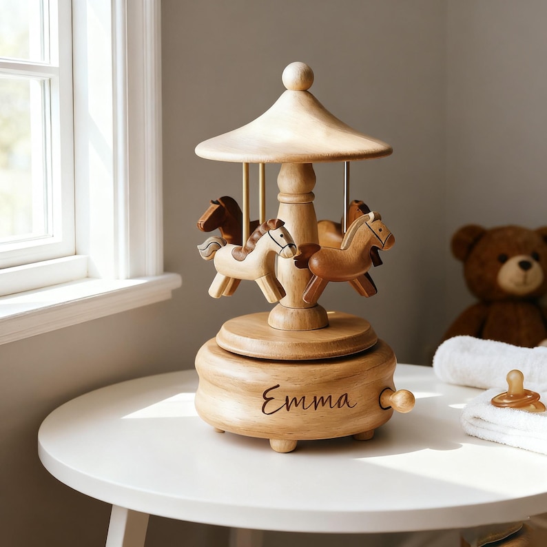 May include: A wooden carousel music box featuring three carved horses in varying shades of brown. The base is circular with a smooth finish and the name Emma is engraved in cursive script on the front. A small wooden knob is attached to the side.