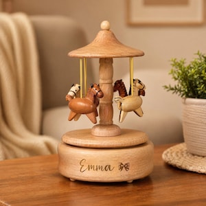May include: A wooden carousel music box featuring a light brown conical roof, a central pillar, and four small carved horses in varying shades of brown and cream suspended by gold-colored rods. The base is engraved with the name Emma and a bow.
