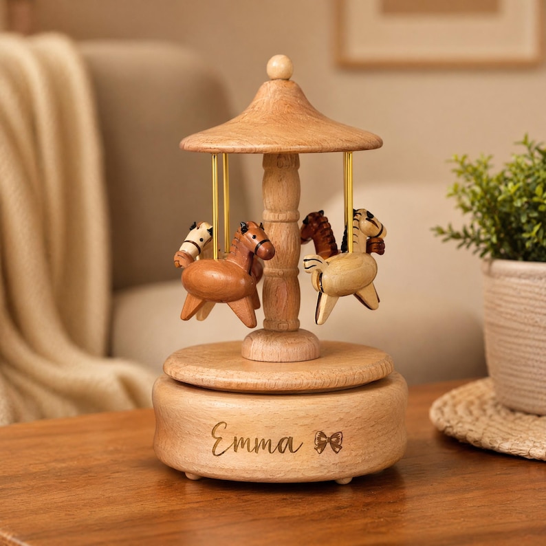 May include: A wooden carousel music box featuring a light brown conical roof, a central pillar, and four small carved horses in varying shades of brown and cream suspended by gold-colored rods. The base is engraved with the name Emma and a bow.