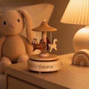 May include: A wooden carousel music box featuring four small horses in varying shades of brown and cream. The circular base is crafted from light-toned wood and features the name Emma printed in a dark script font on the front side.