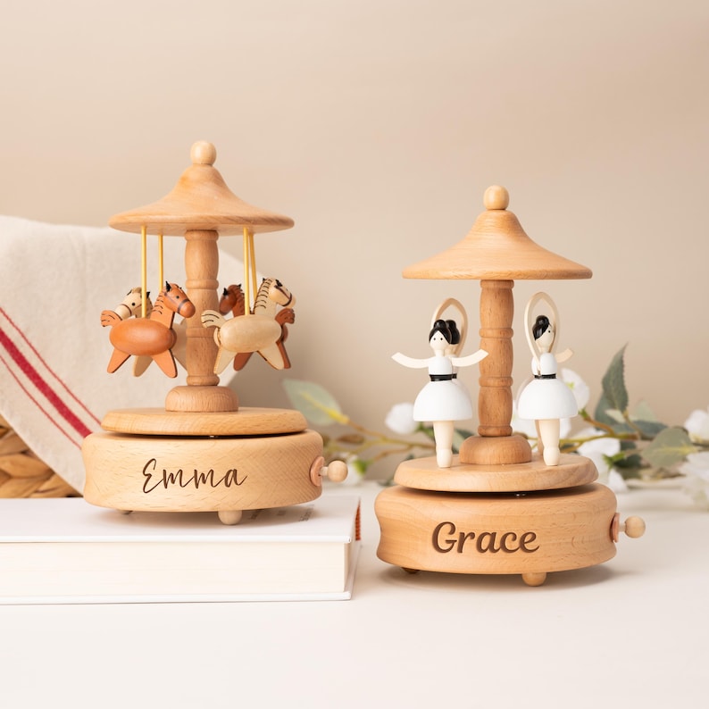 May include: Two wooden carousel music boxes with light wood finish. The left carousel features brown horses and the name Emma engraved on the base. The right carousel features white-clad ballerinas and the name Grace engraved on the base.