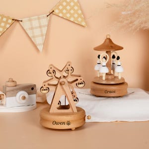 May include: Wooden Ferris wheel music box with the name "Owen" engraved on the base. A wooden carousel music box with ballerina figures is also visible. A wooden camera toy is on the left. A banner with a checkered pattern hangs in the background.