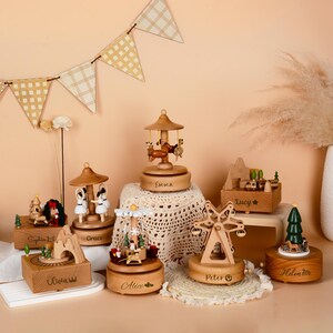 May include: A collection of wooden music boxes with various designs, including a carousel, Ferris wheel, and mountain scene. Each box is a warm brown color and features intricate details and personalized names. The boxes are displayed on a neutral surface with decorative elements.