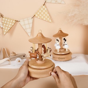May include: Two wooden music boxes with a natural light wood finish. One features miniature brown horses on a rotating carousel, while the other displays white-clad ballerinas. The base of the ballerina music box is engraved with the name Grace.