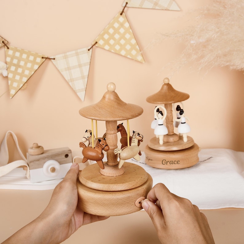 May include: Two wooden music boxes with a natural light wood finish. One features miniature brown horses on a rotating carousel, while the other displays white-clad ballerinas. The base of the ballerina music box is engraved with the name Grace.