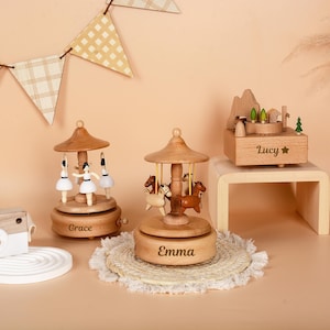 May include: Wooden carousel music boxes with ballerina and horse figurines. The carousel music boxes are engraved with the names "Grace" and "Emma". A wooden music box with a mountain and village scene is also present, engraved with the name "Lucy".
