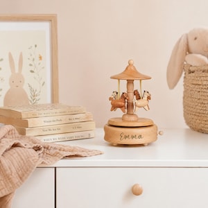May include: A wooden carousel music box featuring four small horses in varying shades of brown and cream. The base is circular with the name Emma engraved in cursive script. A small winding knob is visible on the side of the light-colored wooden base.