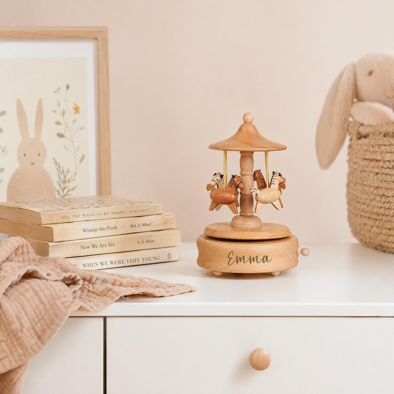 May include: A wooden carousel music box featuring four small horses in varying shades of brown and cream. The base is circular with the name Emma engraved in cursive script. A small winding knob is visible on the side of the light-colored wooden base.
