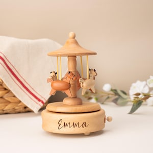 May include: A light-colored wooden carousel music box featuring three small carved horses suspended by thin yellow rods from a central canopy. The circular base of the music box is engraved with the name Emma in a dark, cursive script font.