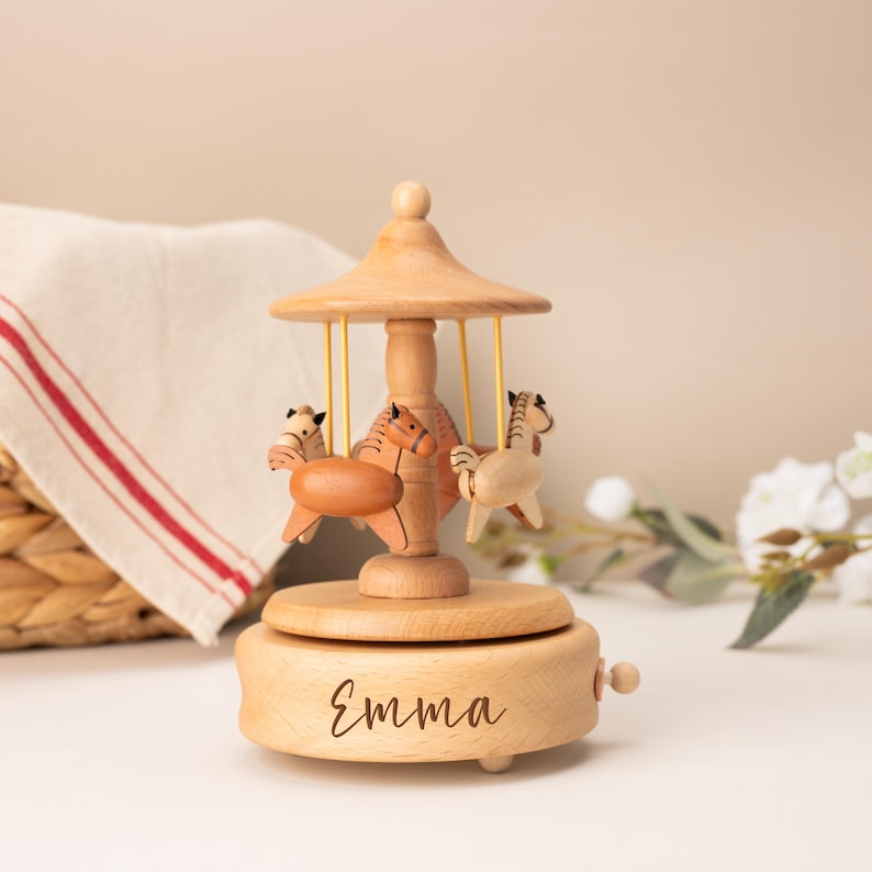 May include: A light-colored wooden carousel music box featuring three small carved horses suspended by thin yellow rods from a central canopy. The circular base of the music box is engraved with the name Emma in a dark, cursive script font.