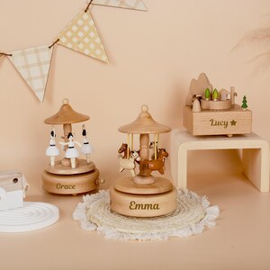 May include: Two wooden carousel music boxes with figures of ballerinas and horses, personalized with the names Grace and Emma, alongside a wooden music box featuring a mountain landscape and the name Lucy engraved on the front.