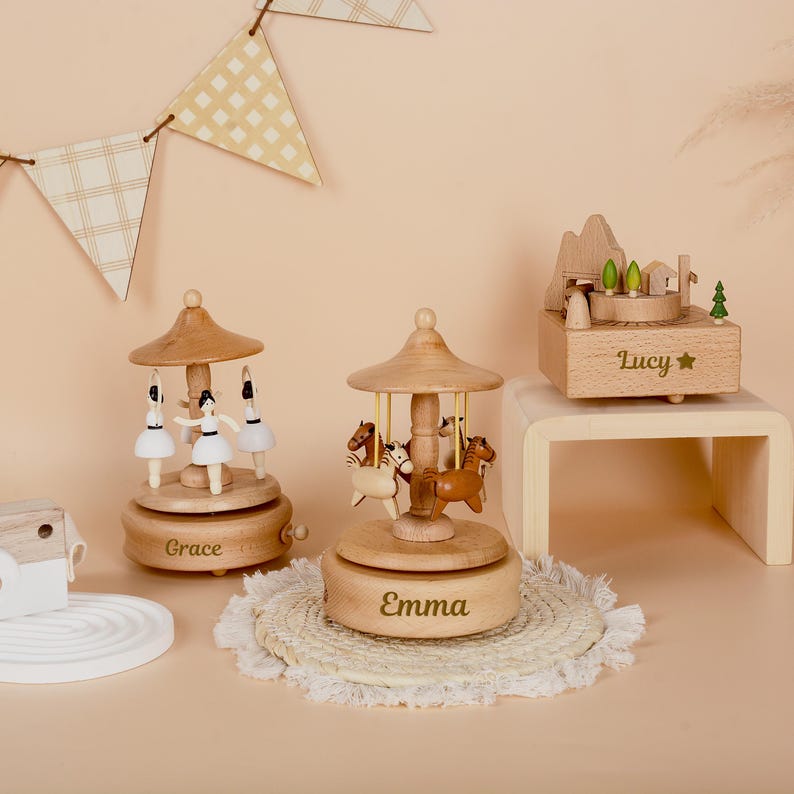 May include: Two wooden carousel music boxes with figures of ballerinas and horses, personalized with the names Grace and Emma, alongside a wooden music box featuring a mountain landscape and the name Lucy engraved on the front.