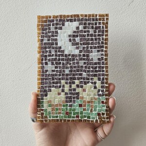 May include: A mosaic art piece depicting a night scene with a moon and grassy area. The artwork is composed of small, square tiles in shades of brown, white, green, and red, framed with a gold border. The mosaic is held in a hand.