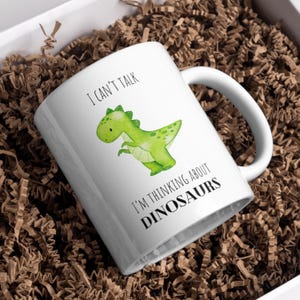 Könnte beinhalten: Weiße Keramiktasse mit einer grünen Dinosaurier-Illustration und dem Text "I CAN'T TALK I'M THINKING ABOUT DINOSAURS". Die Tasse hat einen gebogenen Henkel und steht in einer Box mit braunem Papierfüllmaterial.