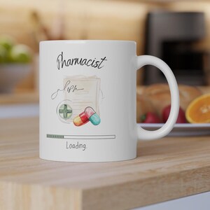 May include: White ceramic mug with the word "Pharmacist" in script, a prescription illustration, and a colorful capsule design. The mug also features a loading bar graphic with the word "Loading" below.