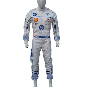 Op de afbeelding: Witte raceoverall met blauwe en rode details, gepresenteerd op een mannequin. De overall heeft sponsorlogo's, waaronder "HP", "Shell" en "UniCredit". Het nummer "16" is zichtbaar op de taille. De overall heeft blauwe strepen.