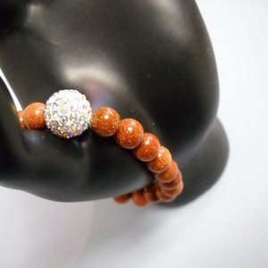 May include: A brown gemstone bracelet with a sparkly silver bead. The bracelet is made of round beads and is on a black hand model.