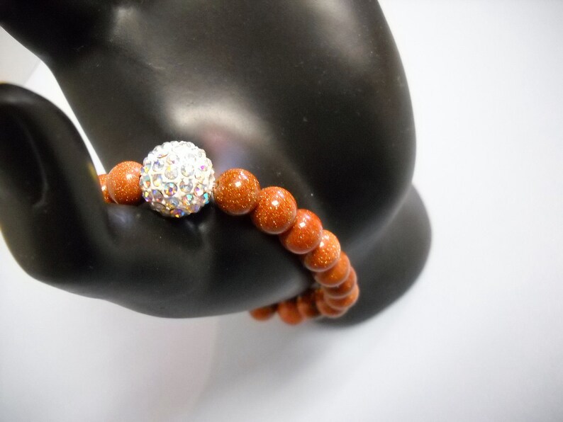 May include: A brown gemstone bracelet with a sparkly silver bead. The bracelet is made of round beads and is on a black hand model.