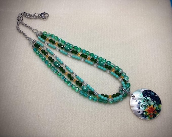 May include: A multi-strand necklace featuring various shades of green and turquoise beads, including faceted and smooth round styles. The necklace is finished with a silver-toned metal chain and a circular pendant decorated with a floral pattern.
