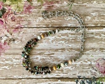 May include: A necklace featuring a silver-toned chain and a cluster of multicoloured, small, rounded beads. The beads display various earthy tones including green, brown, tan, and cream, arranged in a dense, asymmetrical cluster on one side.