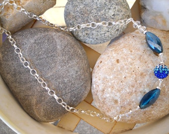 May include: A silver chain necklace with blue and clear crystal beads. The necklace is displayed on a background of grey stones.
