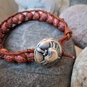 May include: A brown leather bracelet with pink heart-shaped beads and a silver charm featuring a heart with wings and a sword.