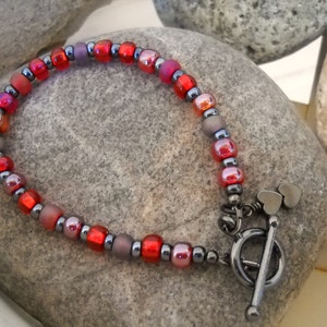 May include: A red, gray, and black beaded bracelet with a heart-shaped clasp. The bracelet is made with small, round beads and has a silver-toned clasp.