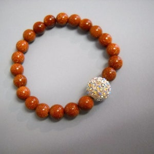 May include: A brown beaded bracelet with a sparkly silver charm. The beads are round and have a speckled texture.