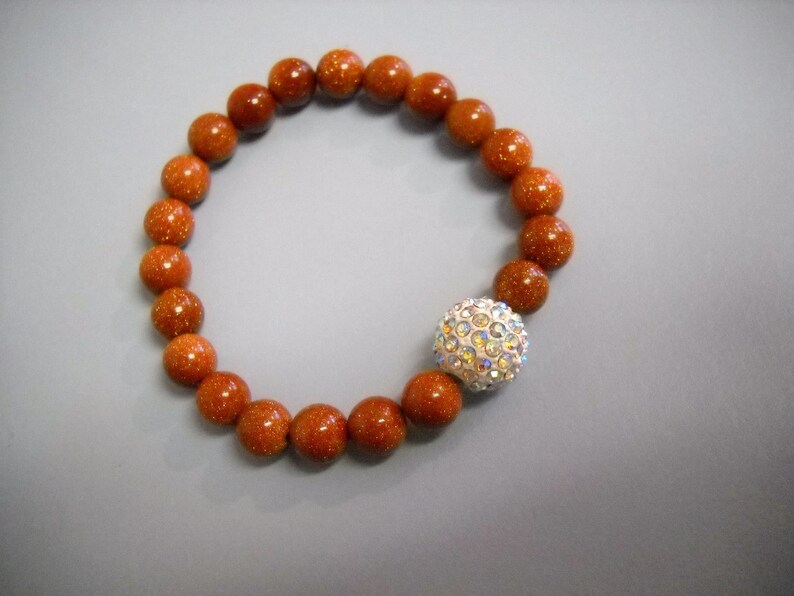 May include: A brown beaded bracelet with a sparkly silver charm. The beads are round and have a speckled texture.