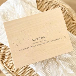 May include: A light-colored wooden box with the name "Matilda" and dates engraved on the surface. The box is decorated with small star-like designs and rests on a woven basket and a white blanket.