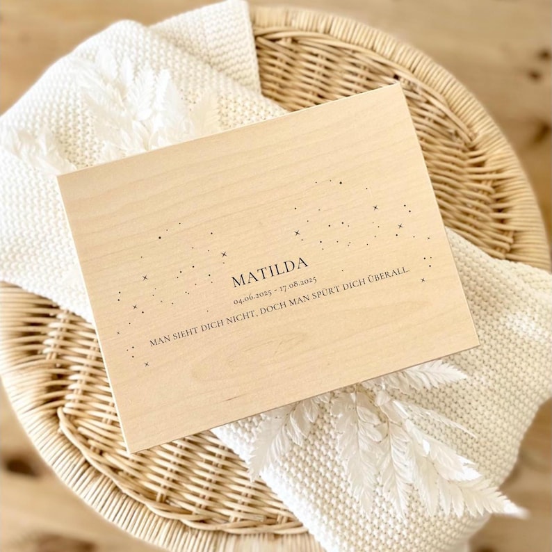May include: A light-colored wooden box with the name "Matilda" and dates engraved on the surface. The box is decorated with small star-like designs and rests on a woven basket and a white blanket.