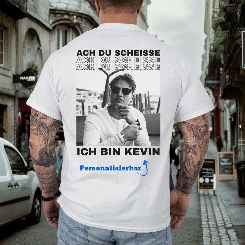May include: White t-shirt with a black and white photo of a man wearing sunglasses. The shirt has the text "ACH DU SCHEISSE" repeated three times above the photo and "ICH BIN KEVIN" below. The word "Personalisierbar" is also printed on the shirt.