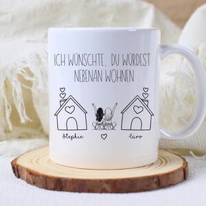 Puede incluir: Taza de cer&aacute;mica blanca con asa, con un dibujo lineal negro de dos casas y dos figuras con los brazos levantados. El texto "ICH W&Uuml;NSCHTE, DU W&Uuml;RDEST NEBENAN WOHNEN" est&aacute; impreso encima de las casas. La taza est&aacute; sobre un posavasos de madera.