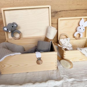 May include: Two open wooden boxes filled with baby items. One box contains a gray elephant teether, a gray blanket, and other items. The other box has a white bunny teether, a brush, and other items. The boxes are on a white surface.