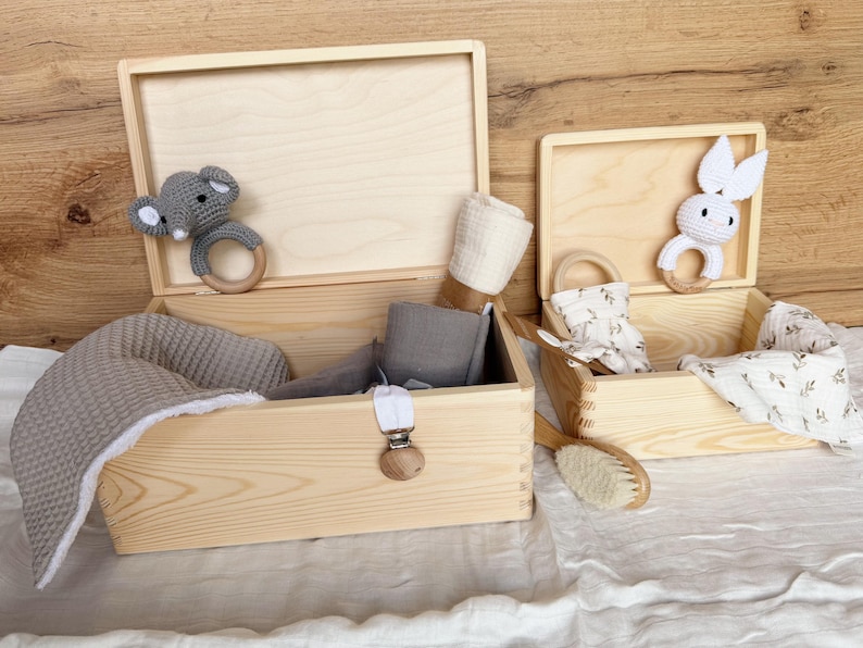 May include: Two open wooden boxes filled with baby items. One box contains a gray elephant teether, a gray blanket, and other items. The other box has a white bunny teether, a brush, and other items. The boxes are on a white surface.