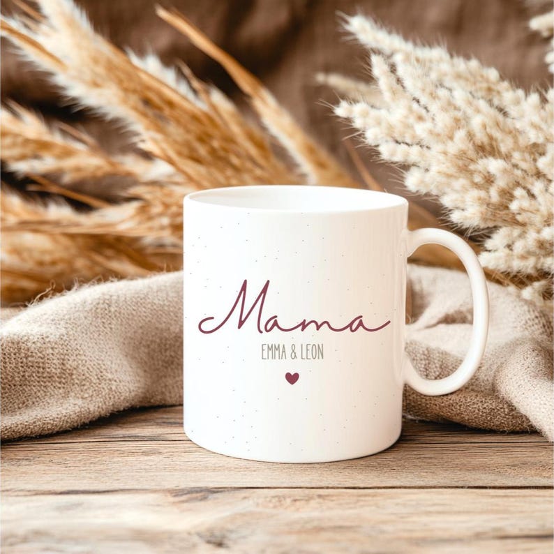 May include: White ceramic mug with the word "Mama" in a cursive font, along with "EMMA & LEON" and a small heart. The mug has a speckled texture and a curved handle. The background includes dried plants and a wooden surface.