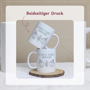 Puede incluir: Dos tazas de cer&aacute;mica blanca apiladas sobre un posavasos de madera. Cada taza presenta un dibujo lineal negro de casas y el texto "ICH WUNSCHTE, DU WURDEST NEBINAN WOHNEN". Las tazas tienen un asa en el lado derecho.