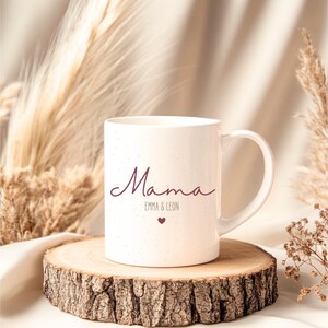 May include: White ceramic mug with the word "Mama" in a cursive font, along with "Emma & Leon" and a small heart. The mug is speckled and sits on a wooden slice, with dried plants in the background.
