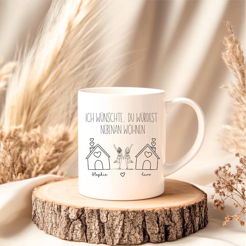 Puede incluir: Taza de cer&aacute;mica blanca con un dibujo lineal negro de dos casas y dos figuras. La taza tiene el texto "ICH W&Uuml;NSCHTE, DU W&Uuml;RDEST NEBENAN WOHNEN" y los nombres "Stehpie" y "Lara". La taza est&aacute; sobre una rodaja de madera.
