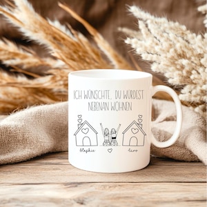 Puede incluir: Taza de cer&aacute;mica blanca con asa. Presenta un dibujo lineal negro de dos casas y dos figuras con los brazos levantados. El texto "ICH W&Uuml;NSCHTE, DU W&Uuml;RDEST NEBENAN WOHNEN" est&aacute; impreso encima de la ilustraci&oacute;n, con los nombres "Stephie" y "Laro" debajo.