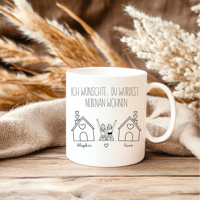 Puede incluir: Taza de cer&aacute;mica blanca con asa. Presenta un dibujo lineal negro de dos casas y dos figuras con los brazos levantados. El texto "ICH W&Uuml;NSCHTE, DU W&Uuml;RDEST NEBENAN WOHNEN" est&aacute; impreso encima de la ilustraci&oacute;n, con los nombres "Stephie" y "Laro" debajo.