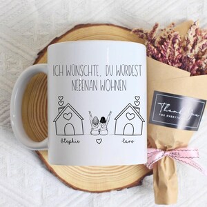 Puede incluir: Taza de cer&aacute;mica blanca con una ilustraci&oacute;n negra de dos casas y dos figuras con los brazos levantados. La taza tiene el texto "ICH W&Uuml;NSCHTE, DU W&Uuml;RDEST NEBENAN WOHNEN" y los nombres "Stephie" y "Laro". Un ramo de flores secas y una tarjeta "Thank You" est&aacute;n al lado.